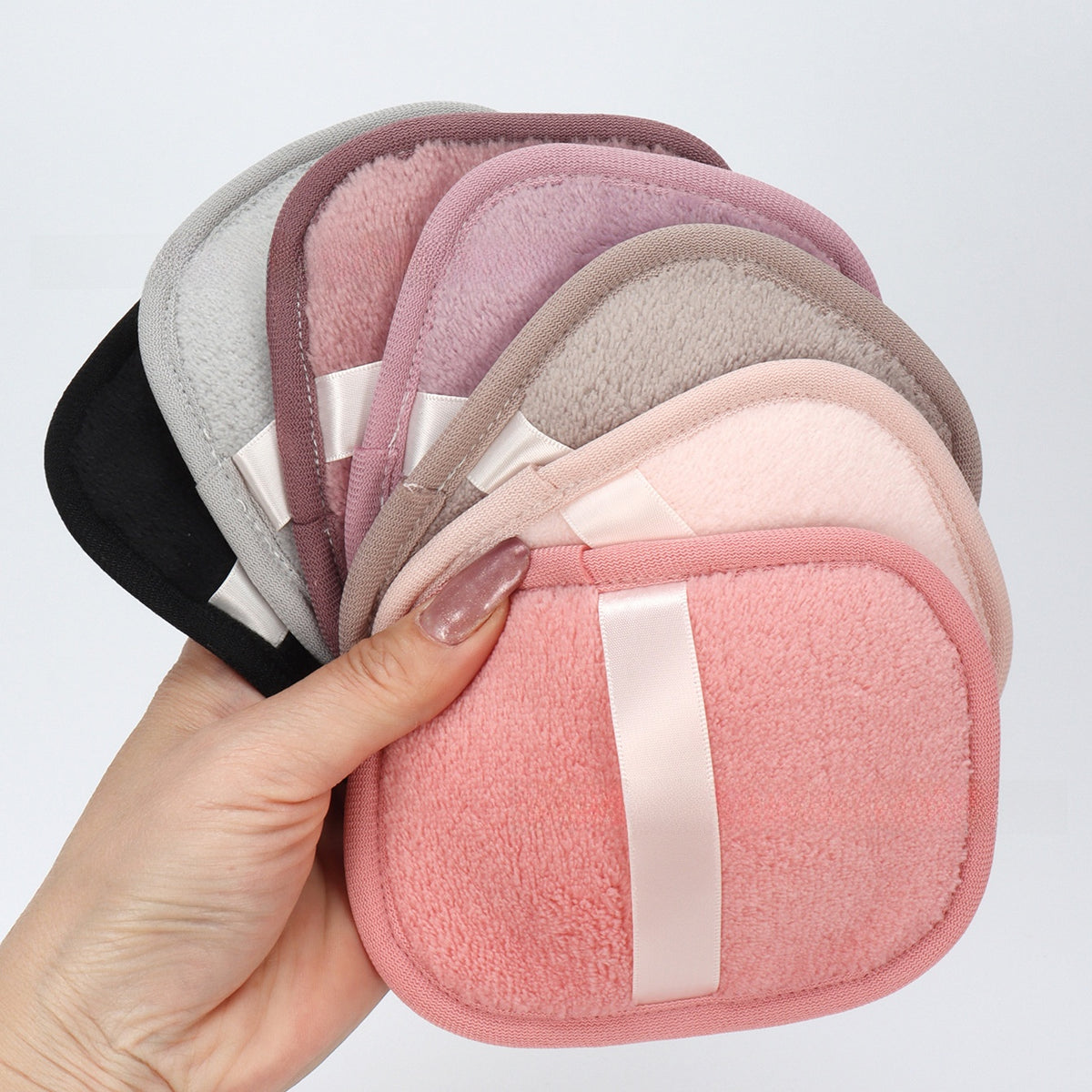 A hand holds a fan of five soft, rounded square makeup sponges in different colors including black, gray, mauve, taupe, and pink, each with a white satin ribbon detail, against a plain light gray background.