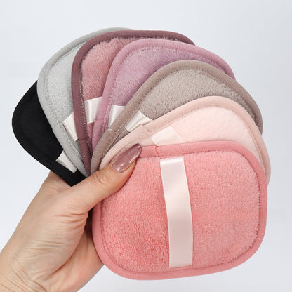 A hand holds a fan of five soft, rounded square makeup sponges in different colors including black, gray, mauve, taupe, and pink, each with a white satin ribbon detail, against a plain light gray background.